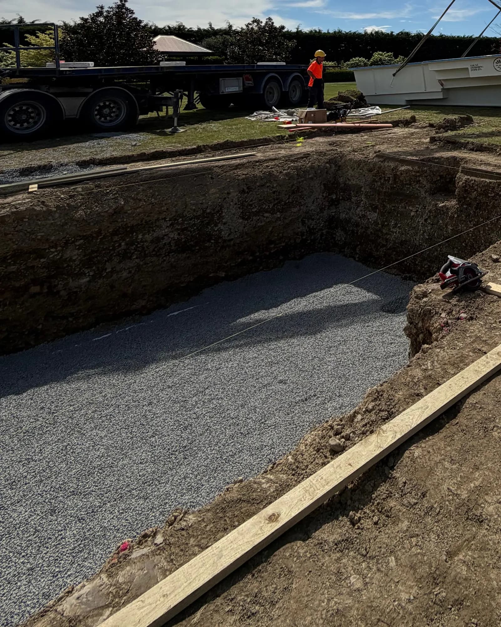 A finished pool dig with a level gravel base, string lines and screed boards, a delivery truck and the shell waiting on the lawn behind