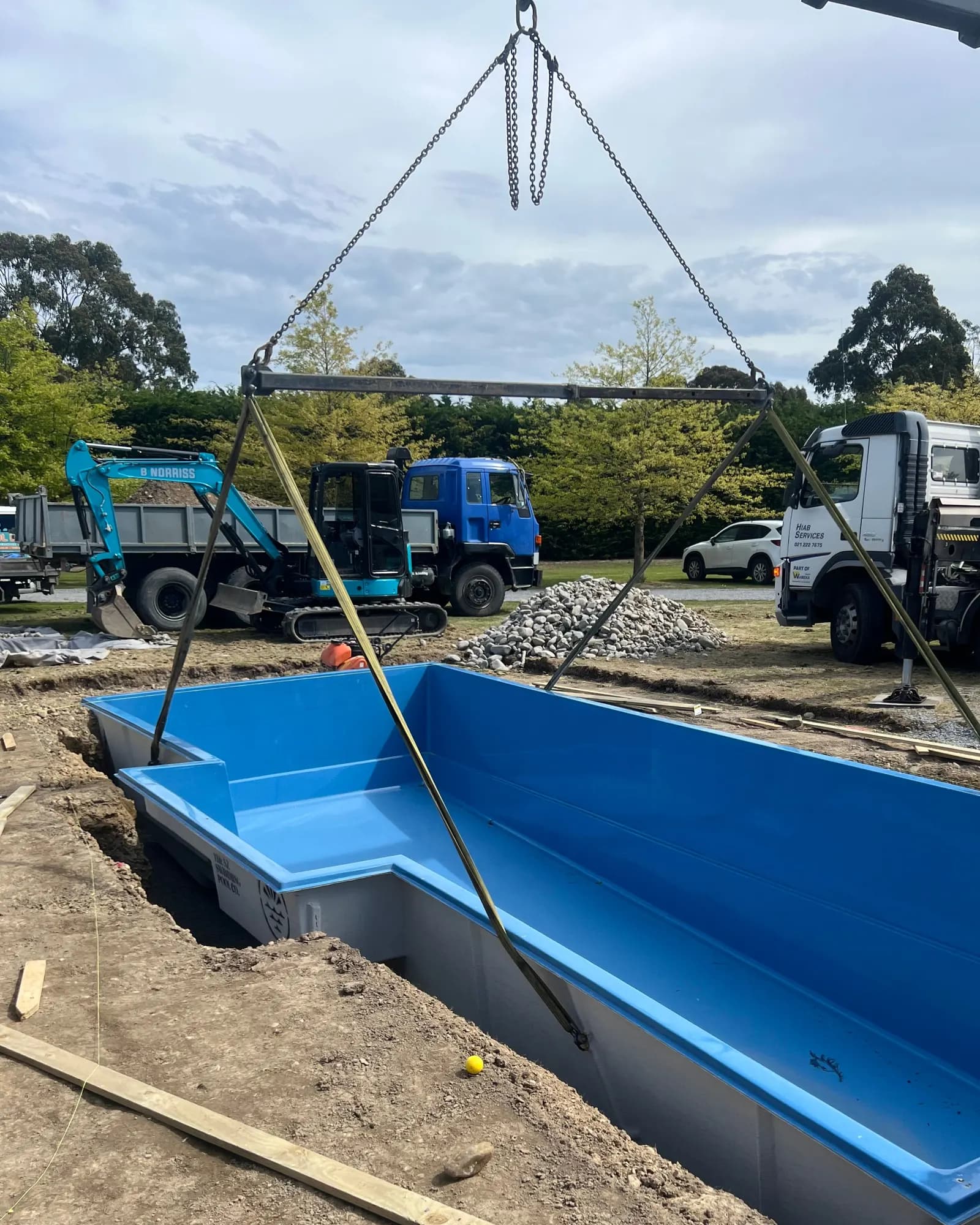 A blue fibreglass shell hangs from crane chains as it is lowered into the dig, a digger and trucks parked behind