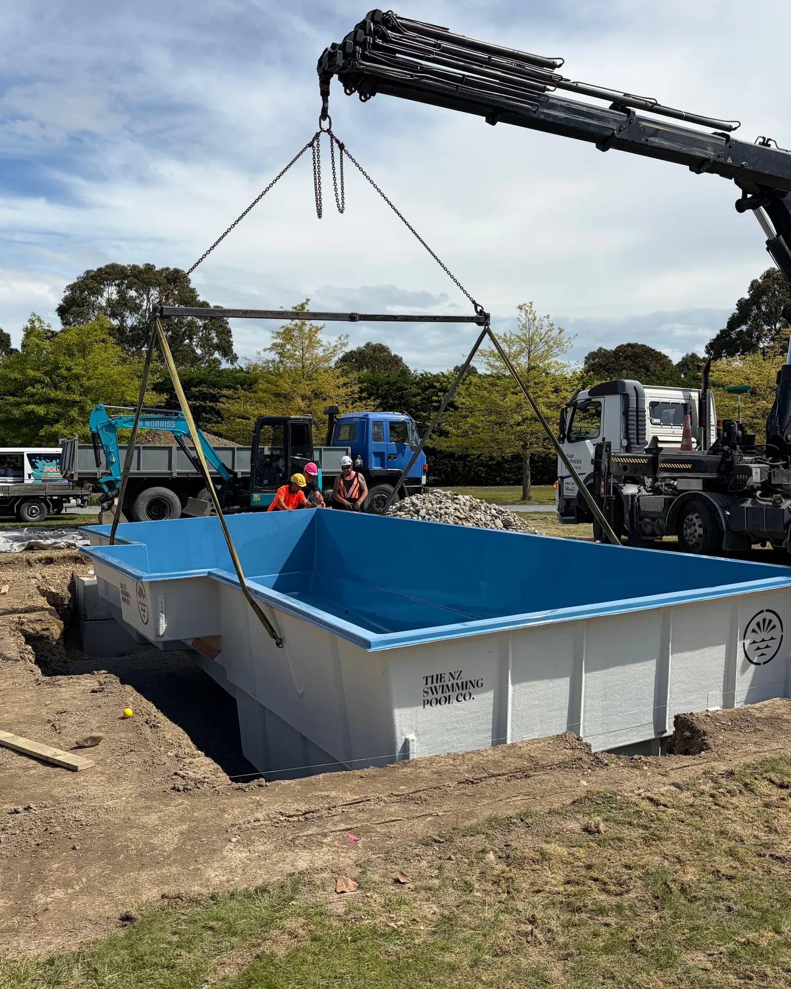 A truck-mounted crane holds a blue fibreglass shell on its spreader bar as it sits down into the dig