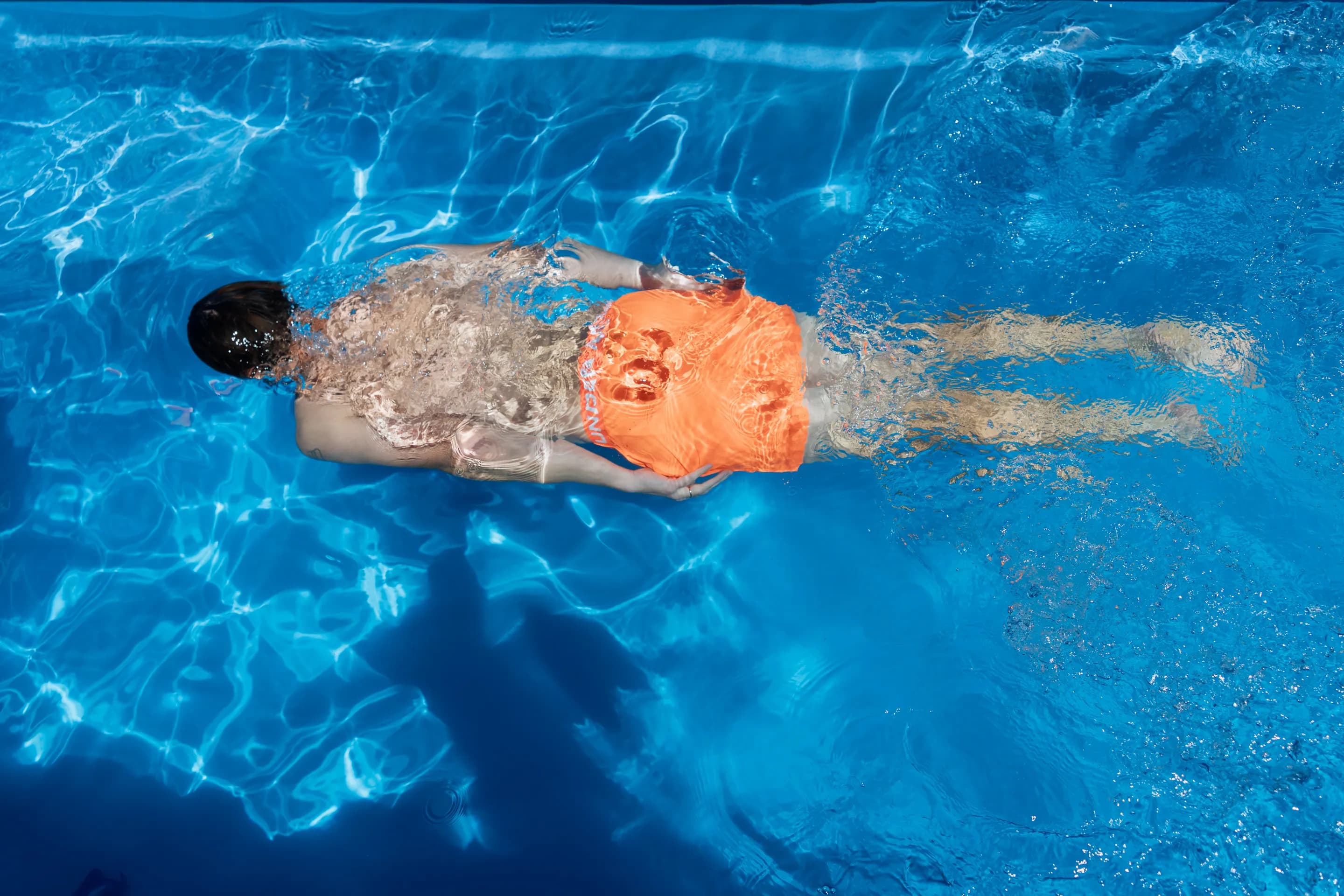 A swimmer in orange shorts glides underwater through a bright blue fibreglass pool